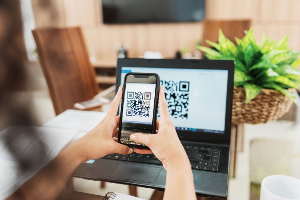 Mãos de uma pessoa posicionando o celular para ler um QR Code na tela de um notebook