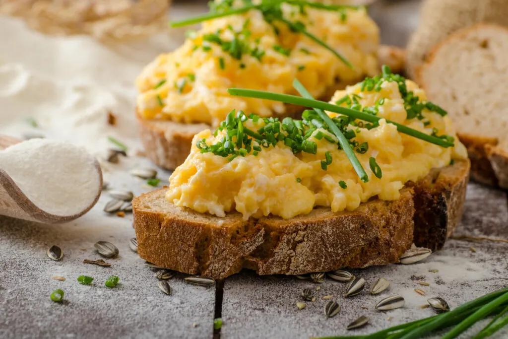 Dois pedaços de pães com ovo mexido e cebolinha picada em cima de uma mesa de madeira