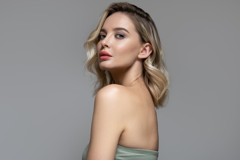 Mulher com cabelo loiro e raiz escura de lado, com top cinza e em fundo cinza