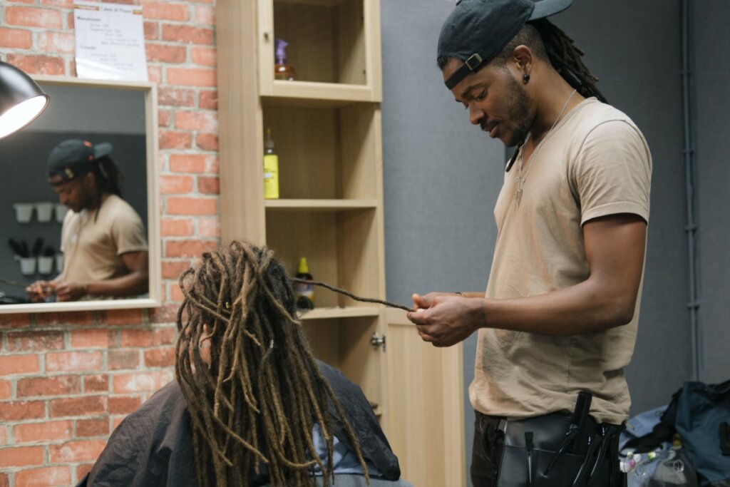 Homem com cabelo dreads no cabelo usando boné preto, camisa bege e calça jeans fazendo dreads em pessoa sentada, em salão de beleza