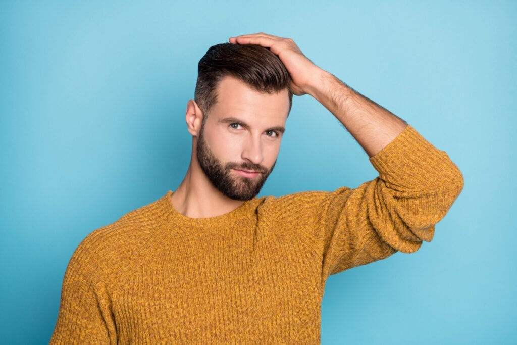 Homem com cabelo em topete, curto e usando barba. Com um suéter ocre em fundo azul-turquesa