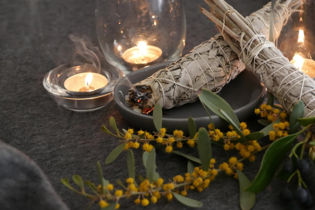 Bastão de sálvia em prato cinza com velas e ramos de flores amarelas ao lado