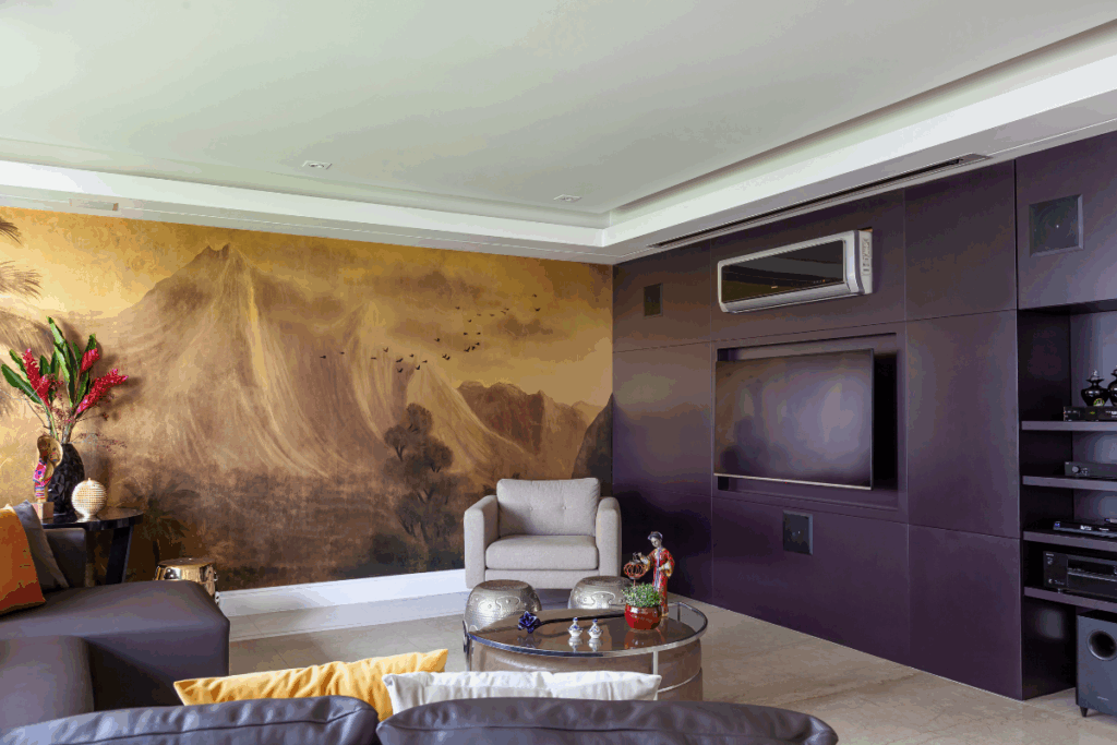 Sala de estar com parede escura com TV e ar condicionado e outra parede com papel de parede de uma paisagem de montanha em tons de bege e sofá marrom e poltrona cinza e mesa de centro em vidro com objetos de decoração em cima