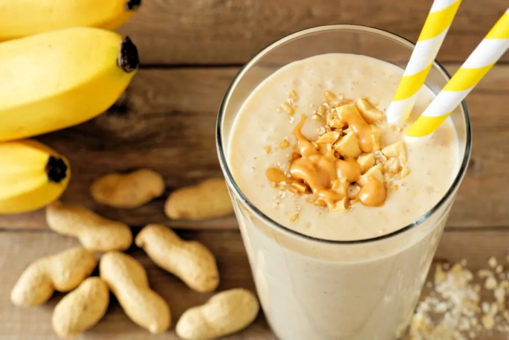 Copo de vidro com vitamina de amendoim e banana, decorada com pedaços de amendoim e mel, com um canudo amarelo e branco 