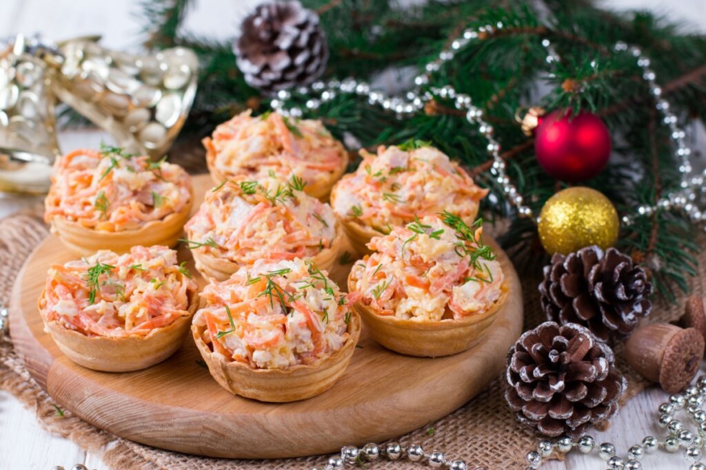 Tábuas com tartaletes preenchidas com salada de frango, cenoura e ovos, finalizadas com dill picado, ao lado de enfeites de Natal