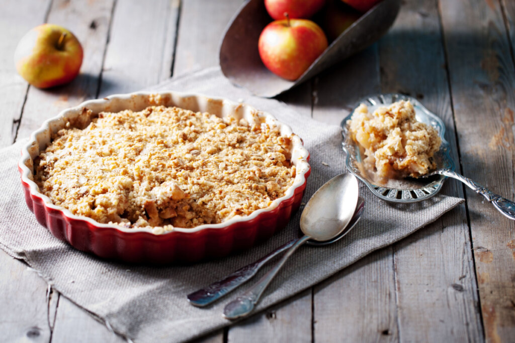 Forma vermelha com crumble de maçã em cima de uma mesa de madeira e ao lado de maçãs