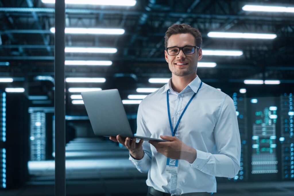 Homem com cabelo curto, usando óculos preto e camisa de botões branca com notebook na mão em ambiente com computadores