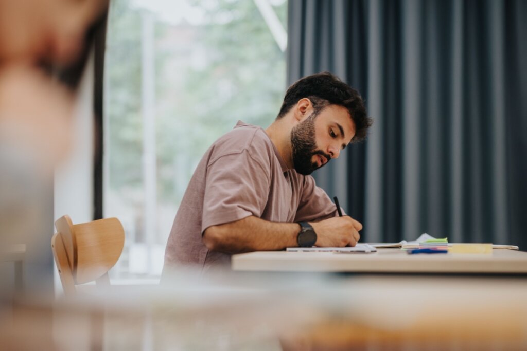 Estudante com o cabelo preto e barba usando camiseta rosa escrevendo