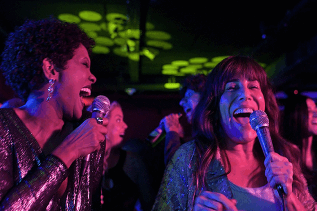 Imagem de duas mulheres segurando microfones e cantando em um ambiente escuro com luzes coloridas e outras pessoas ao fundo