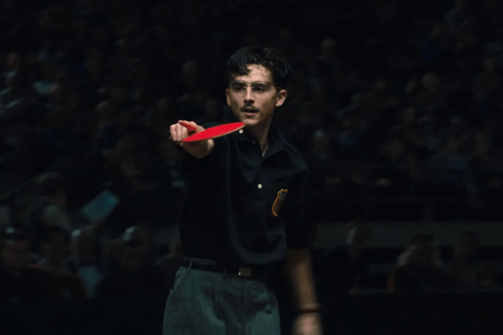 Personagem de Timothée Chalamet segurando uma raquete de tênis de mesa em posição de ataque durante uma partida, em um ambiente fechado, com plateia desfocada ao fundo