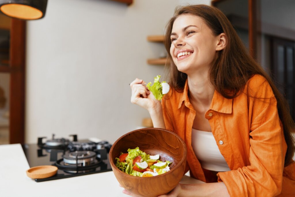 Mulher com cabelo liso, solto, usando regata branca e camisa de botões laranja com uma tigela de madeira com salada em uma mão e um garfo com salada na outra, encostada em balcão na cozinha
