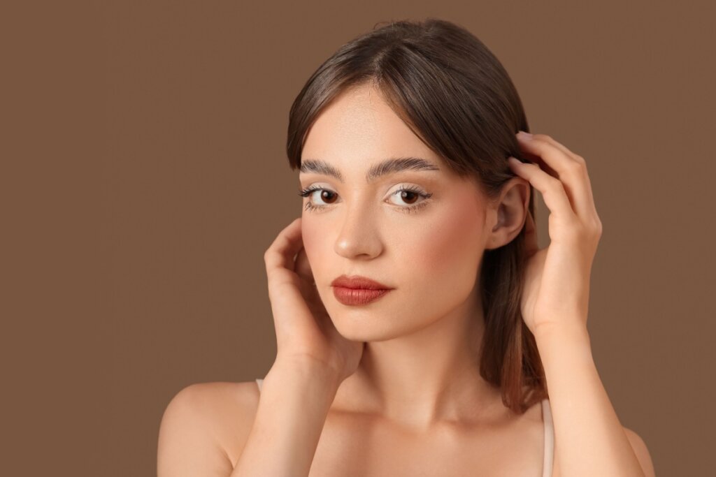 Mulher com maquiagem em tons quentes, blush marcado, pele uniforme e lábios rosados, tocando o cabelo diante de fundo marrom liso