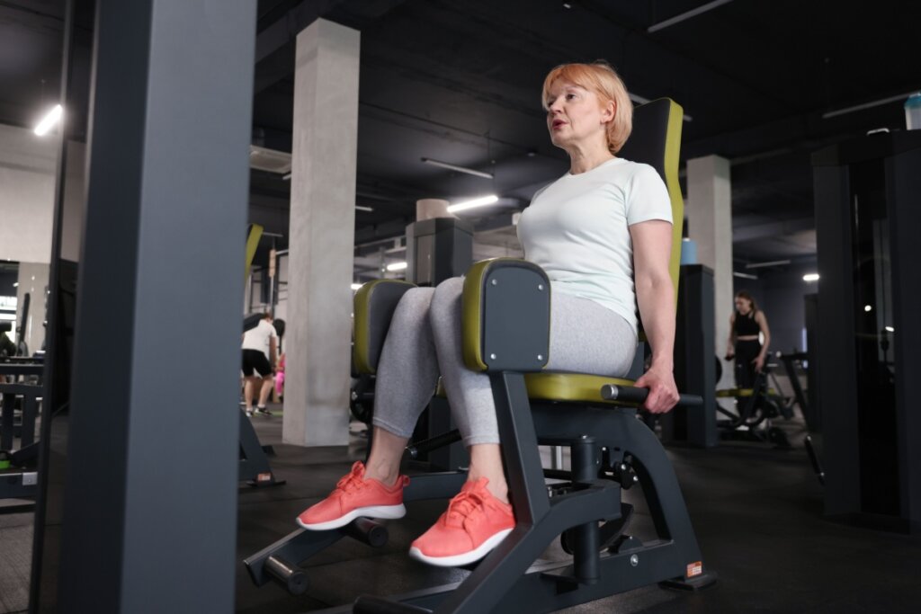 Mulher mais velha com cabelo curto loiro, usando camiseta branca, calça cinza e tênis rosa fazendo musculação na academia