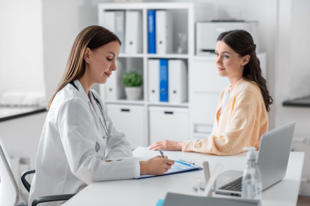 Mulher com cabelo preto, preso parcialmente usando camisa de manga longa laranja em consulta com medica que tem o cabelo liso, solto e está usando jaleco branco anotando informações em prancheta