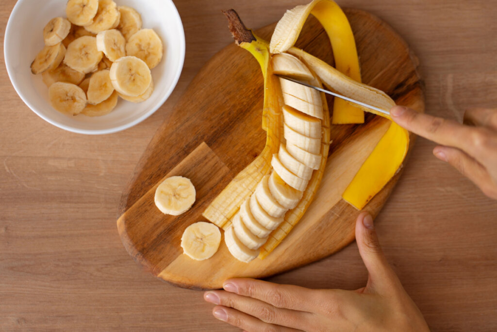 Vista superior de mãos femininas cortando uma banana madura em uma tábua de madeira clara. A banana está parcialmente descascada, com metade já fatiada em rodelas uniformes sobre a casca. Ao lado esquerdo, há uma tigela branca pequena contendo várias rodelas de banana já cortadas