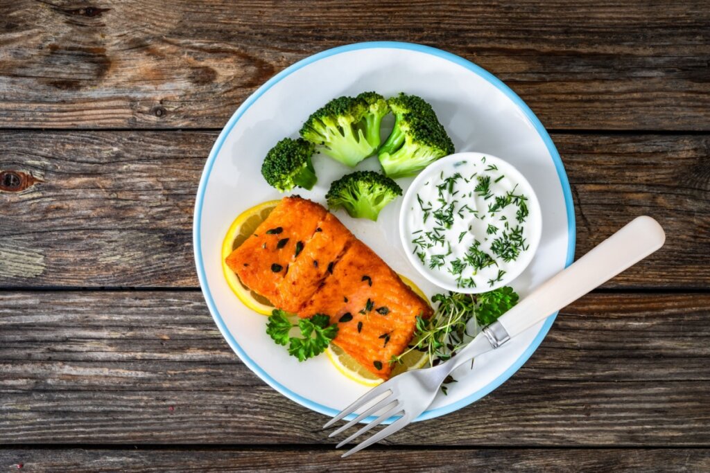 Um prato branco com borda azul contendo um filé de salmão grelhado, temperado com ervas e servido sobre fatias de limão siciliano. Ao lado, há pequenos buquês de brócolis cozidos no vapor e uma tigela pequena com molho de iogurte de ervas frescas. O prato está sobre uma mesa de madeira rústica, acompanhado por um garfo.