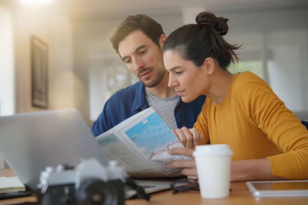 Casal olhando mapa e computador, mulher com cabelo preso em coque usando camiseta amarela e homem com cabelo curto, barba, usando camiseta cinza e camisa jeans