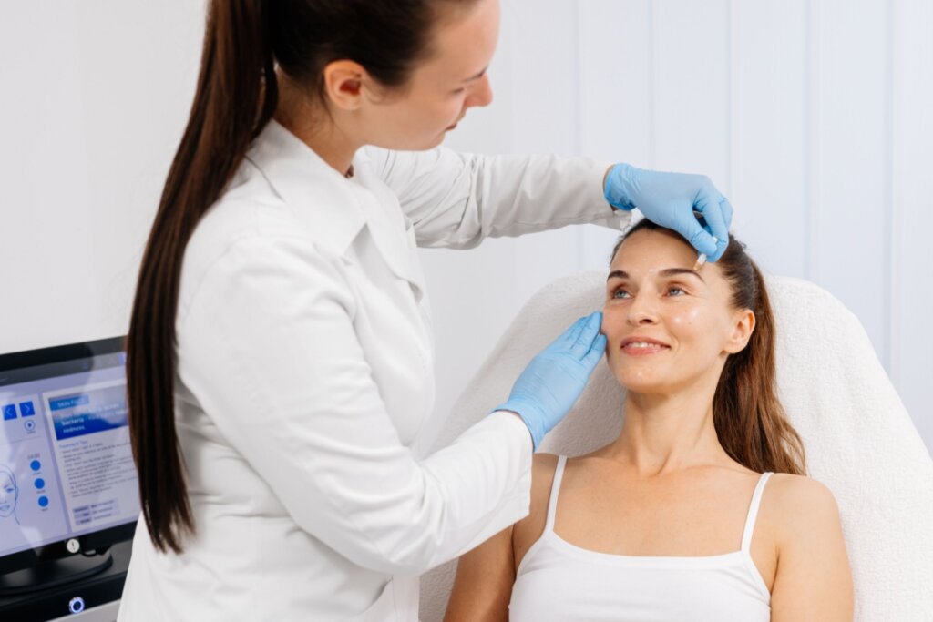 Médica com cabelo preso em rabo de cavalo, usando jaleco branco e luvas azuis, analisando o rosto de paciente sentada em maca usando regata branca