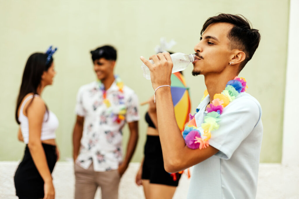 Um homem jovem de perfil e usando um colar de flores coloridas, bebendo água de uma garrafa plástica transparente durante o carnaval. Ao fundo, em um plano desfocado, outras três pessoas conversam em um ambiente ensolarado com uma parede verde clara.