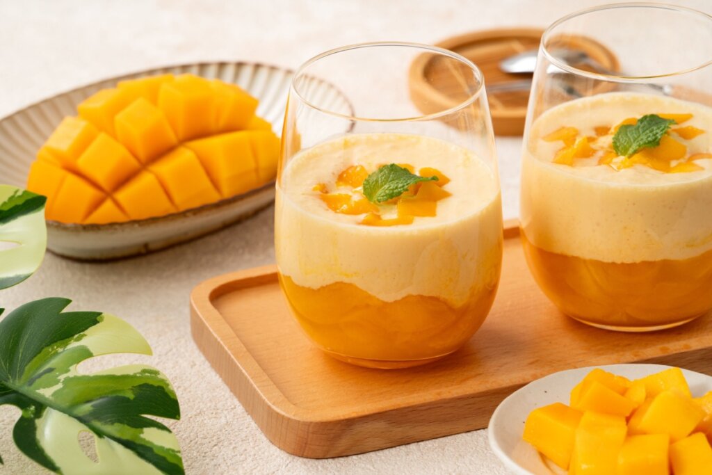 Taças com sobremesa de manga em camadas, com base de creme de manga, cobertura clara e finalização com cubos da fruta e folha de hortelã