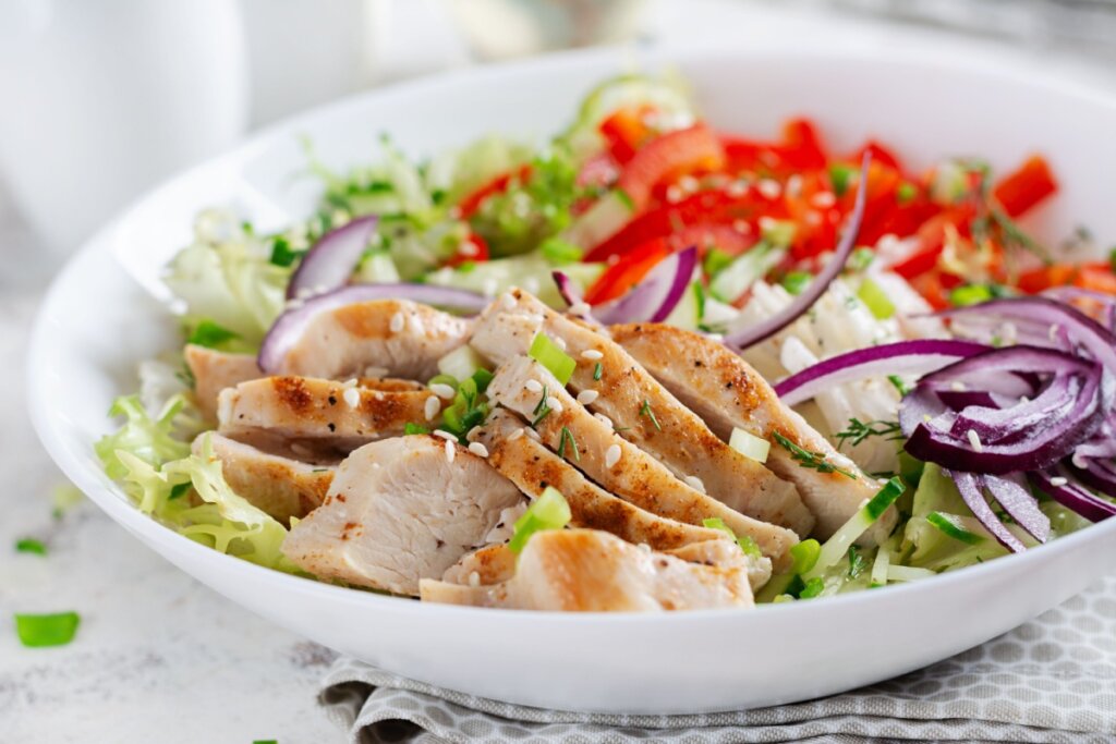 Salada morna de frango grelhado com vegetais servido em tigela branca de cerâmica em cima de bancada