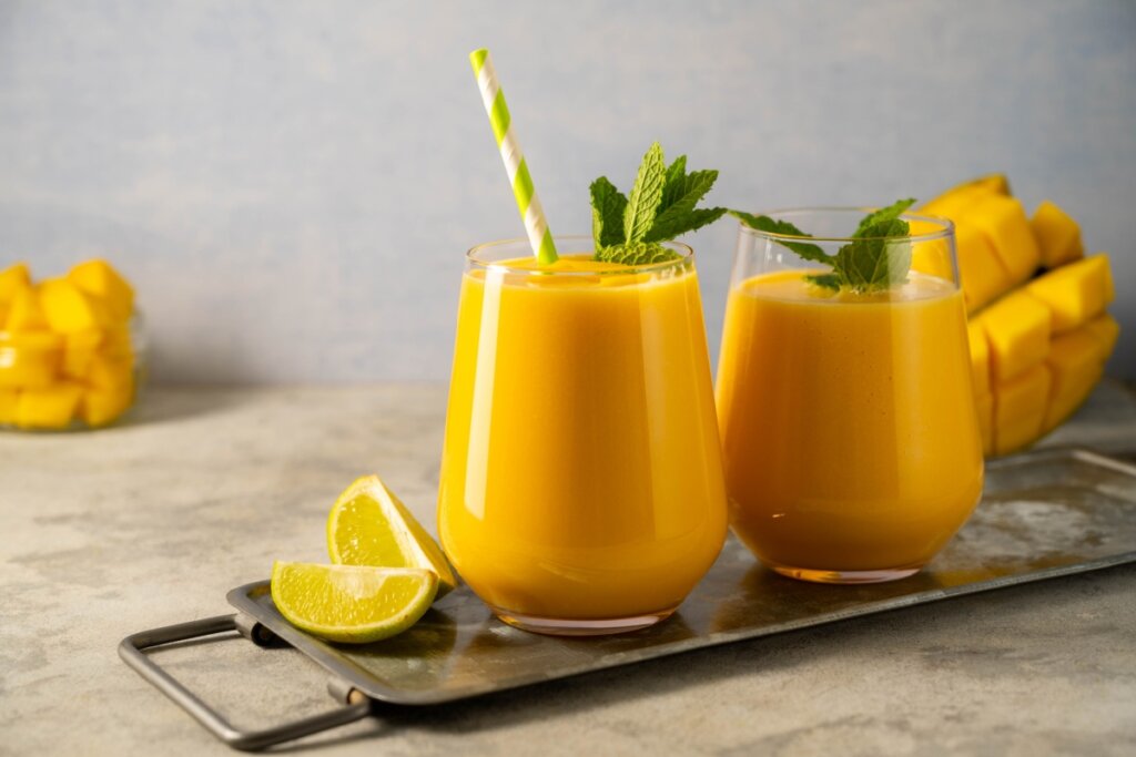 dois copos de vidro com vitamina amarela com hortelã em cima sobre bandeja de metal com fatias de limão ao lado e manga ao fundo