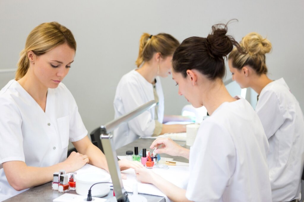 Fotografia colorida mostrando quatro mulheres em um curso de manicure. Elas estão organizadas em uma bancada cinza, vestindo jalecos brancos profissionais. A mulher em destaque, no primeiro plano à esquerda, observa o trabalho enquanto a colega à sua frente manipula esmaltes coloridos e ferramentas de manicure sobre uma toalha branca. O ambiente é claro, limpo e bem iluminado por luminárias de mesa articuladas, transmitindo uma atmosfera de aprendizado técnico e profissional.