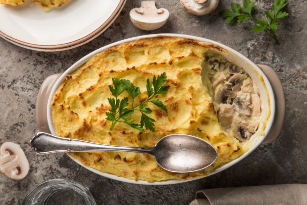 Escondidinho de mandioca com frango e cogumelos em travessa oval, com superfície dourada marcada e interior cremoso exposto