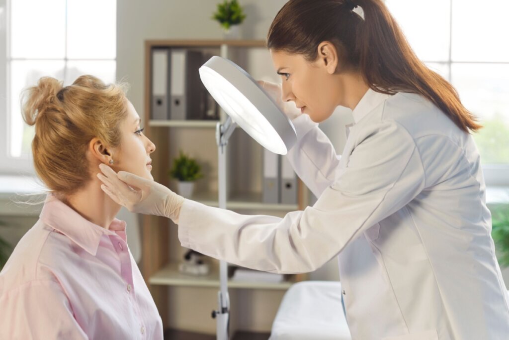 Dermatologista analisando pele do rosto de paciente em consultório, a médica está com o cabelo preso em rabo de cavalo, usando jaleco e luvas brancas e a paciente tem o cabelo loiro preso em coque e está usando camisa social rosa