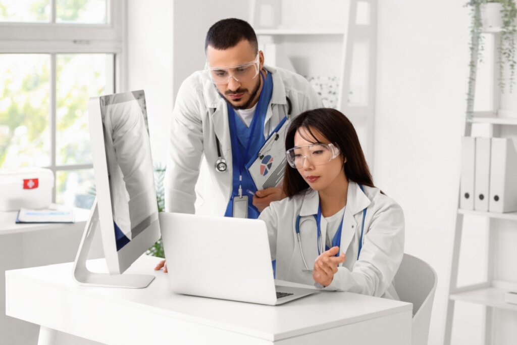 Um homem e uma mulher usando uniforme de médico e óculos de proteção analisando dados no computador em laboratório