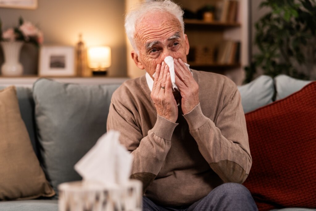 Homem idoso, sentado em um sofá, assoando o nariz com um papel 