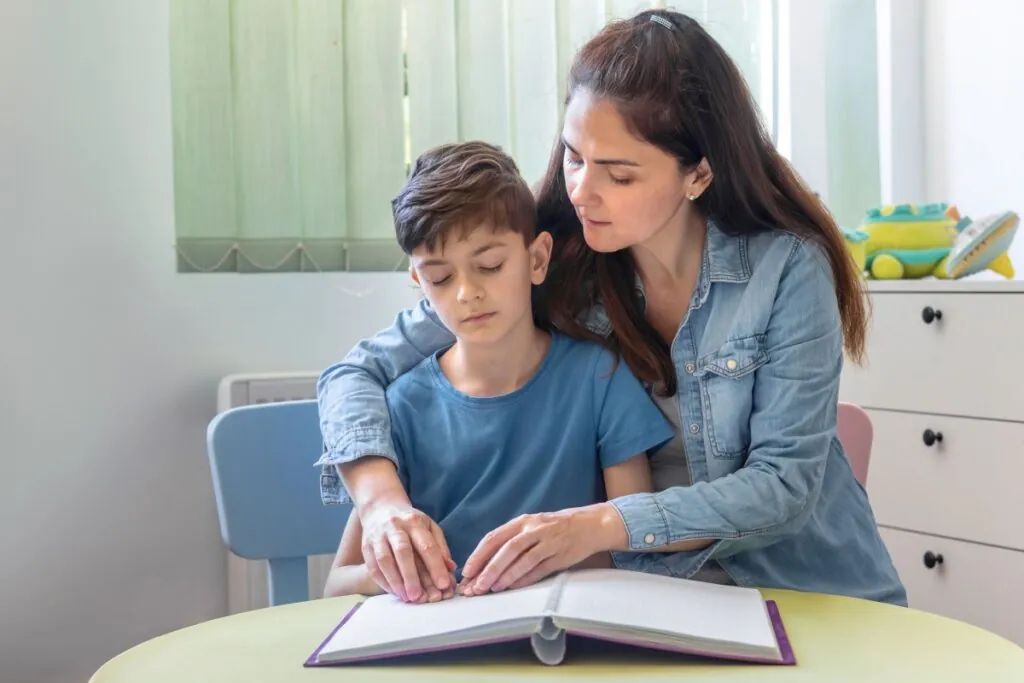 Uma mulher sentada ao lado de um menino, os dois com as mãos sobre um livro de Braille aberto em uma mesa. A mulher veste uma jaqueta jeans e tem o braço esquerdo em volta dos ombros do menino. O menino veste uma camiseta azul e tem os olhos fechados. Seus dedos estão sobre as páginas em relevo do livro de Braille, com as pontas tocando os pontos. O livro é roxo com páginas brancas e espirais no meio. A mesa é redonda com tampo verde claro. Atrás deles, há uma cadeira azul claro e um móvel de gavetas branco. O fundo é iluminado por luz natural de uma janela com cortinas verticais verde claro.