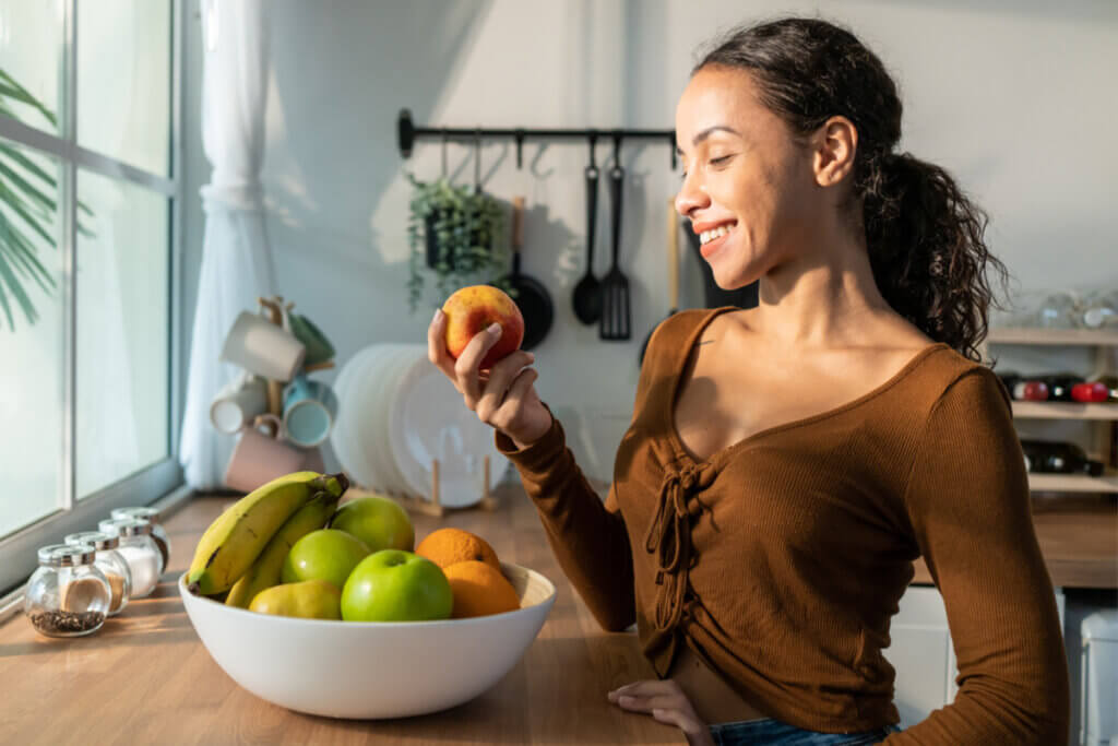 Uma mulher jovem de pele parda e cabelos cacheados sorri enquanto segura uma maçã vermelha em uma cozinha iluminada pelo sol. Ela veste uma blusa marrom de manga longa e está posicionada à frente de um balcão de madeira. Sobre o balcão, há uma fruteira branca generosa contendo bananas, maçãs verdes e laranjas. Ao fundo, vê-se uma janela com luz natural, utensílios de cozinha pendurados e prateleiras organizadas, criando uma atmosfera leve e saudável.