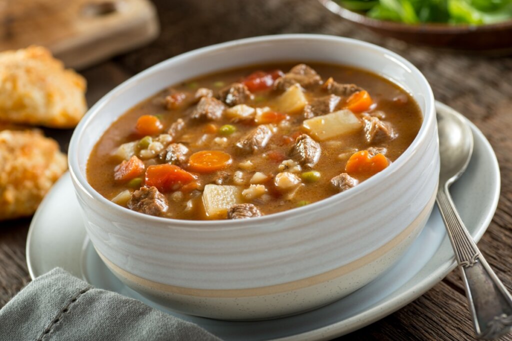 Sopa de carne com cevada e legumes em tigela de cores claras em cima de prato de mesma tonalidade