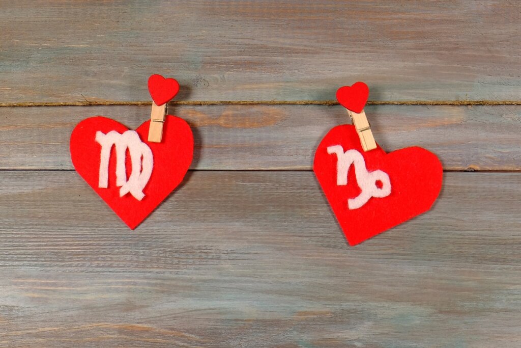 Dois corações de feltro vermelho presos por prendedores em um varal de corda. À esquerda, o símbolo de Virgem em branco; à direita, o símbolo de Capricórnio. O fundo é composto por tábuas de madeira acinzentadas.