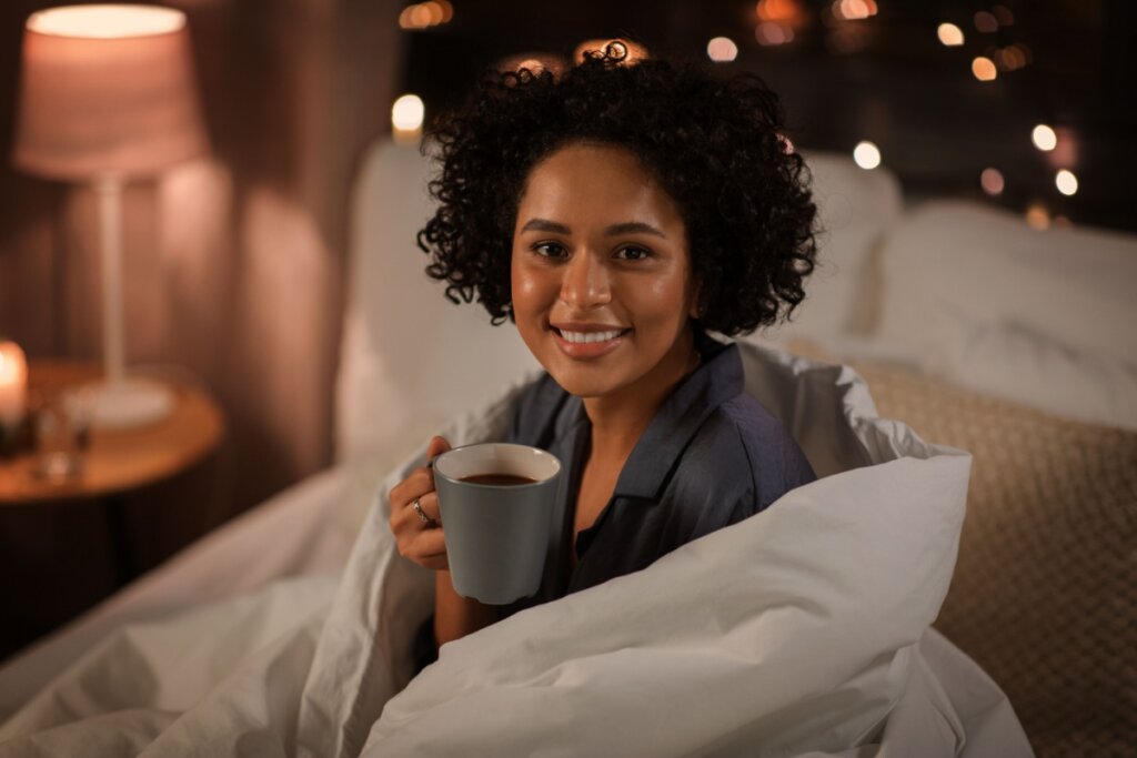 Jovem com cabelo curto cacheado, usando pijama cinza, enrolada em coberta tomando chá, sentada na cama antes de dormir