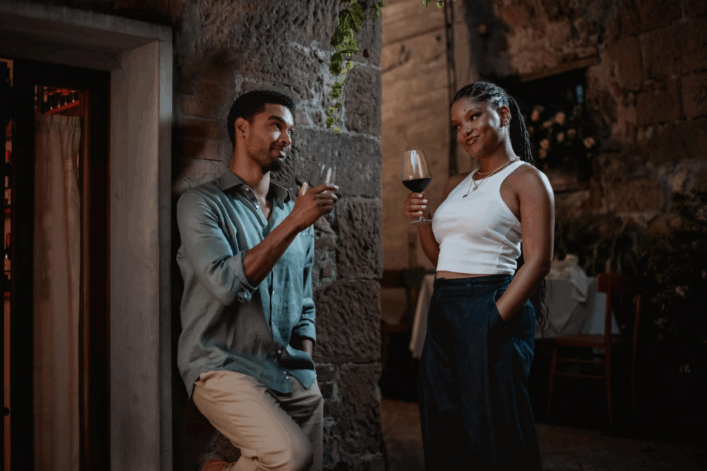 Imagem de um casal bebendo uma taça de vinho encostados no muro de uma casa, o homem tem o cabelo curto e está usando camisa verde e calça bege e a mulher está com o cabelo preso, usando regata branca e calça preta