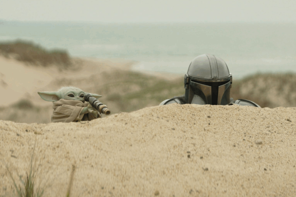 Imagem de um homem usando um capacete de metal e com um personagem verde, usando uma roupa bege espionando atrás de uma duna de areia