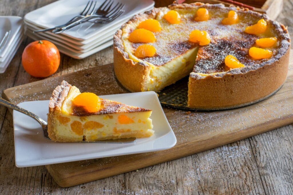 Uma fatia de cheesecake de tangerina em primeiro plano sobre uma pequena travessa branca, servida com uma espátula de metal. Ao fundo, o restante da torta redonda sobre uma tábua de madeira, decorada com gomos de tangerina e polvilhada com açúcar de confeiteiro. A torta possui uma base de massa alta e dourada, com um recheio cremoso e levemente chamuscado no topo. À esquerda, uma tangerina inteira e uma pilha de pratos brancos com garfos completam a cena rústica sobre uma mesa de madeira.
