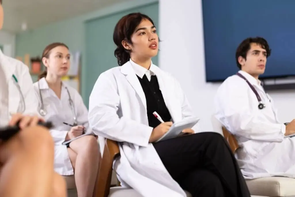 Em uma sala de aula ou auditório, uma jovem estudante de medicina com cabelos escuros presos está sentada, olhando atentamente para frente, como se assistisse a uma palestra. Ela veste jaleco branco sobre uma blusa preta e camisa social, e segura um bloco de notas e uma caneta. Ao seu redor, outros estudantes de jaleco também prestam atenção na aula, com uma tela de TV preta visível ao fundo.