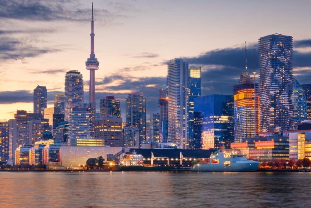 Uma vista do horizonte da cidade de Toronto ao entardecer, com a Torre CN proeminente e um navio cargueiro atracado na orla do Lago Ontário.