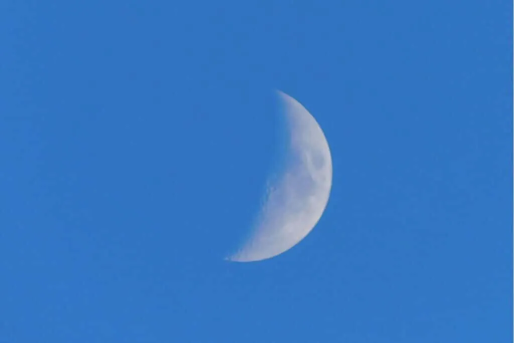 Uma fotografia centralizada, de plano médio, de uma lua minguante contra um céu claro e azul durante o dia.
