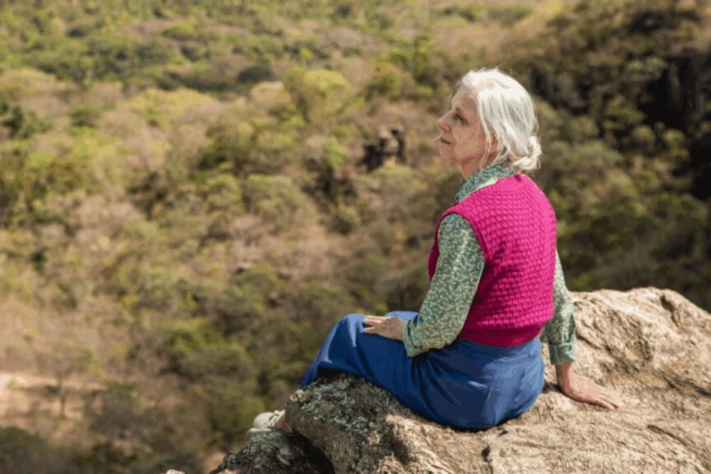 Uma mulher idosa de cabelos brancos presos e blusa de tricô rosa está sentada sobre uma grande rocha no alto de um morro, observando a paisagem com vegetação ao fundo.