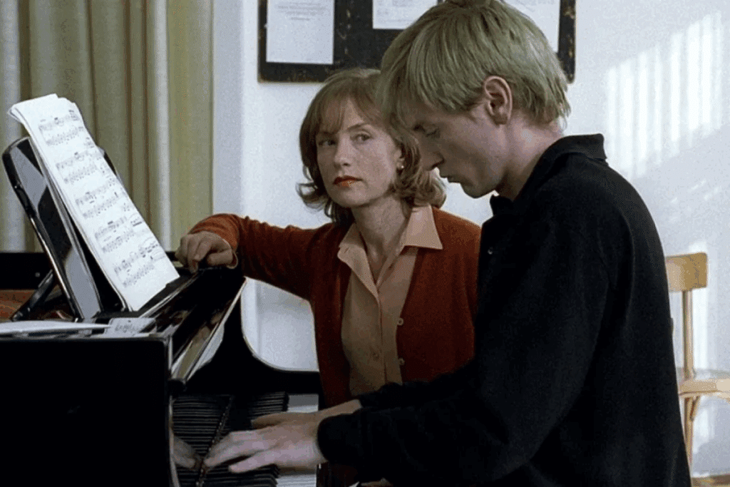 Mulher com cabelo curto, usando camisa rosa-clara e blusa de lã vermelha sentada em banco de piano olhando para jovem tocando piano, ele tem o cabelo curto, loiro e está usando casaco preto