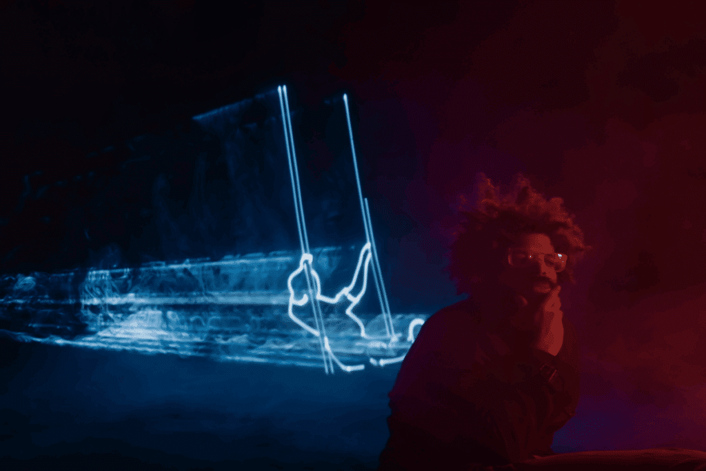 Imagem de um homem com o cabelo cacheado curto, usando óculos com o rosto apoiado na mão em fundo preto com vermelho e com figuras iluminadas em azul no fundo