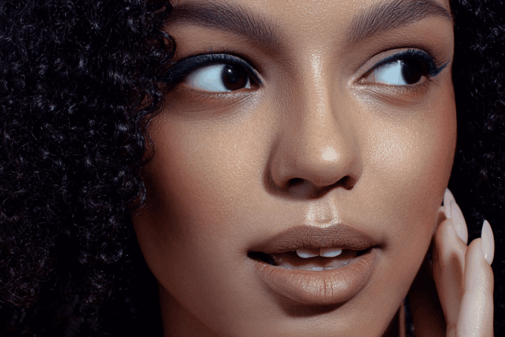 A imagem mostra um close-up do rosto de uma mulher com pele uniforme e iluminada, destacando uma maquiagem sofisticada em tons neutros. Os olhos recebem delineado e sombra azul intensa, enquanto os lábios aparecem com batom marrom de acabamento fosco