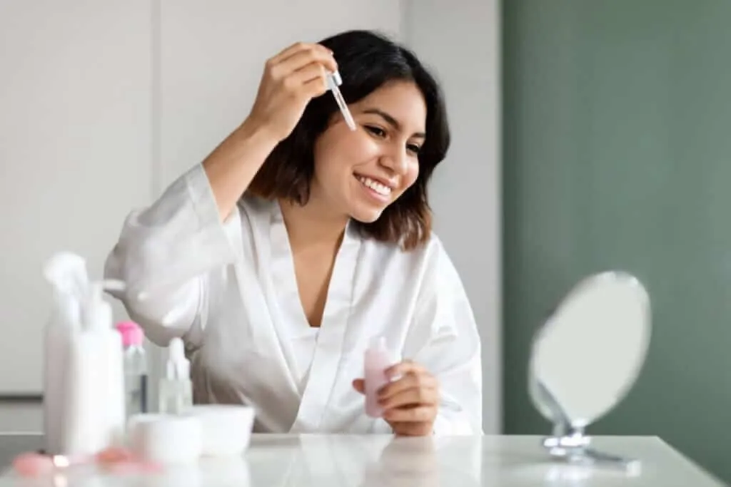 A imagem mostra uma mulher realizando uma rotina de cuidados com a pele diante do espelho. Ela está vestindo um roupão branco e segurando um frasco com conta-gotas, aparentemente aplicando ou prestes a aplicar um sérum facial.