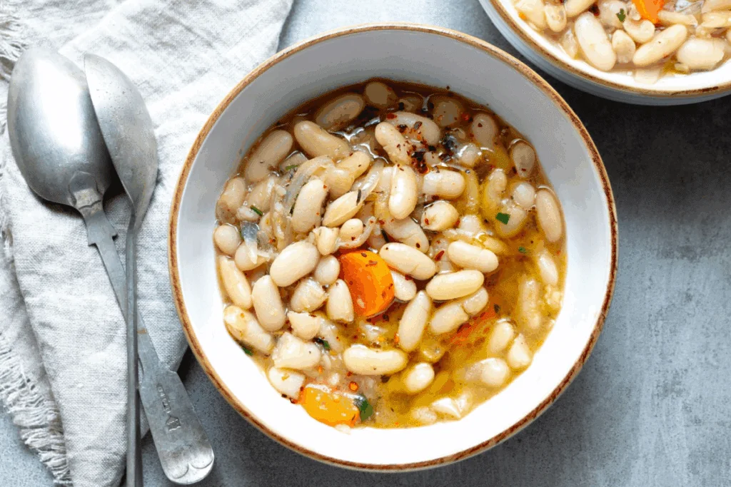 Bowl com sopa de feijão-branco e cenoura em uma mesa cinza. Ao lado, colher e pano branco