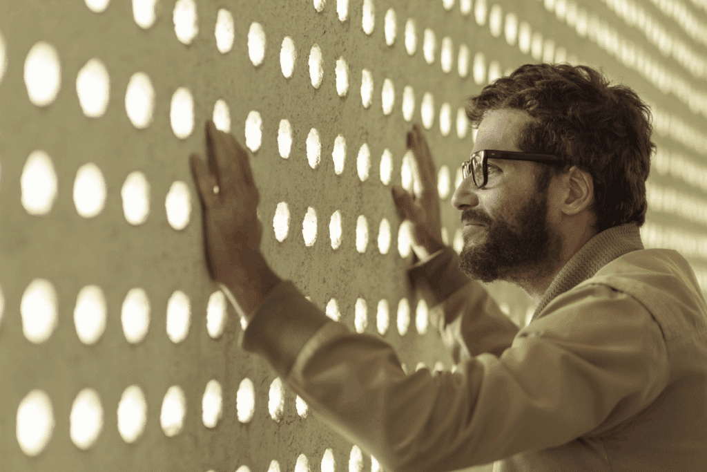 Fotografia em plano médio de um homem de perfil com barba, óculos de armação escura e jaqueta bege, apoiando as duas mãos sobre uma parede com furos redondos vazados por onde passa a luz do sol.