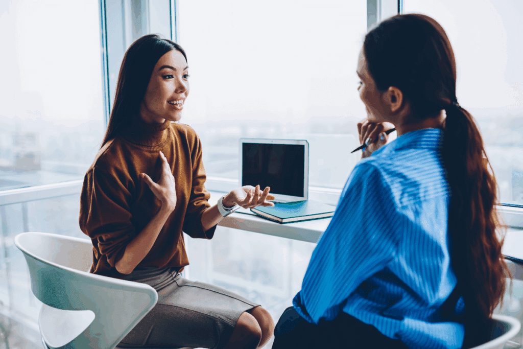 Duas mulheres conversando no trabalho, uma tem o cabelo liso, longo e está usando blusa marrom e saia cinza e a outra está de costas com o cabelo preso e usando camisa azul 